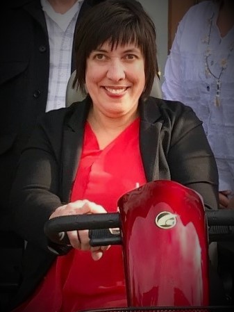 Image of Nicole Bohn smiling, wearing a red shirt and black blazer sitting in her power wheelchair.